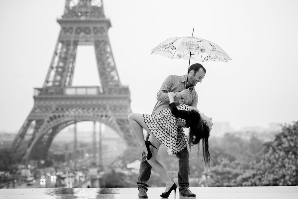 A man holds a woman in a dance-like pose under an umbrella in front of the Eiffel Tower, symbolizing love and romance in a picturesque Paris setting.